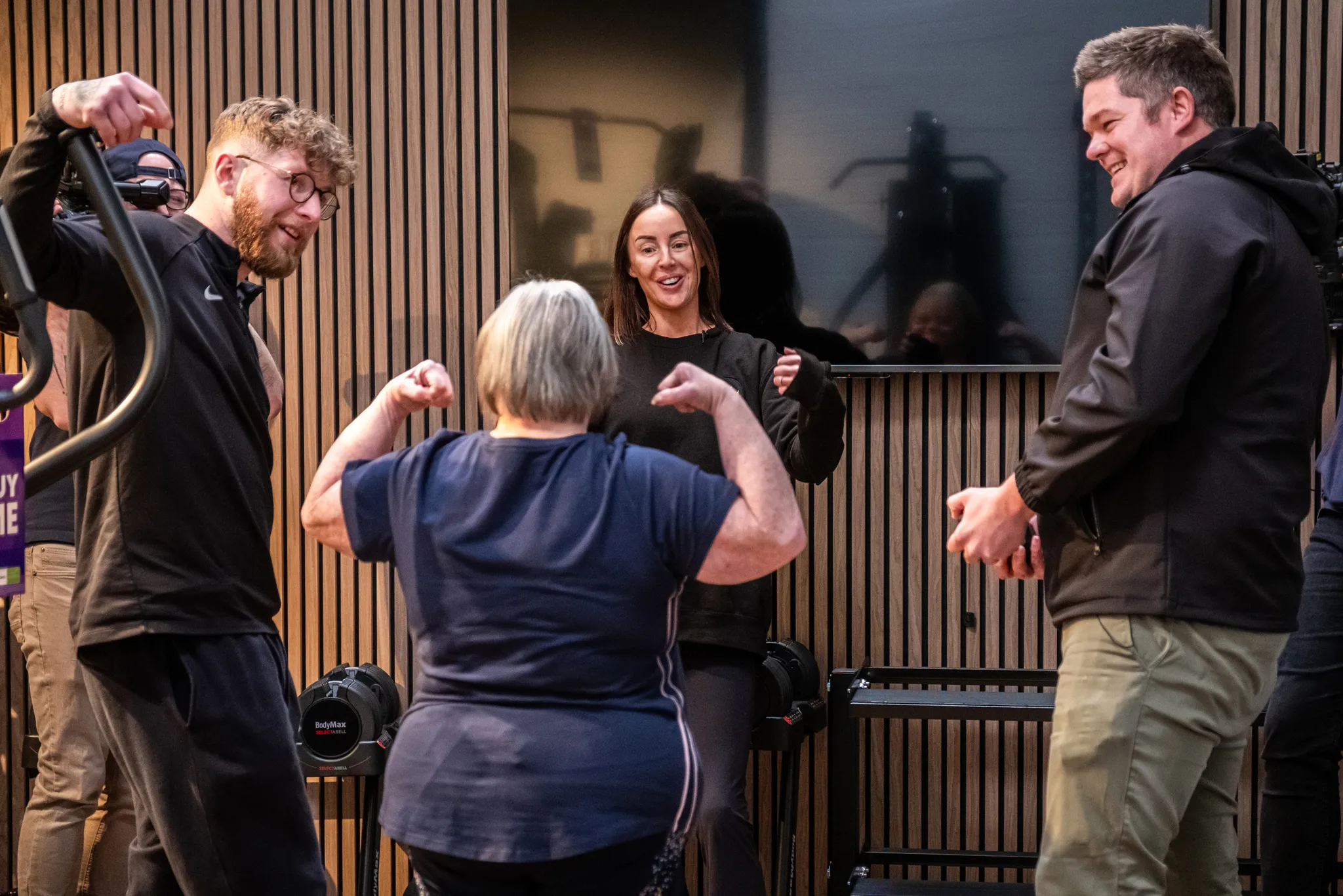 A woman with Down's syndrome seen from behind flexing her muscles surrounded by laughing and smiling gym staff and trainers in a modern fitness studio with wooden slat wall panelling, with a photographer visible in the background